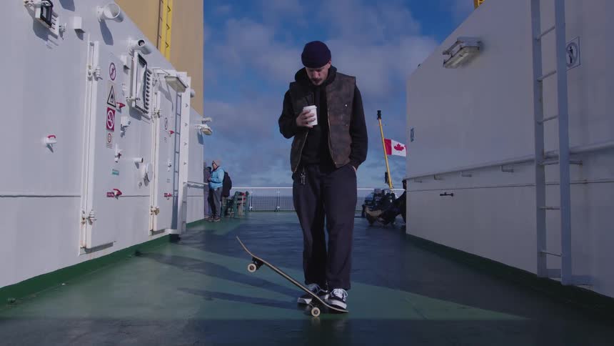 The video features a man standing on what appears to be the deck of a large cargo ship or ferry holding a white cup and taking a sip he is dressed in dark clothing including a camopatterned vest and a beanie and is standing in front of a rais