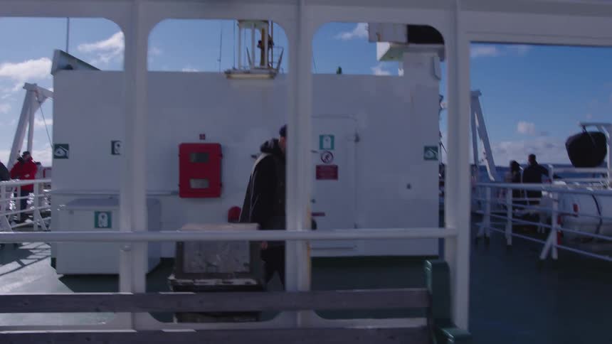 A man wearing a dark beanie and dark jacket walks with a skateboard and a cup across a white boat deck passing a large white wall and other passengers under a sunny blue sky