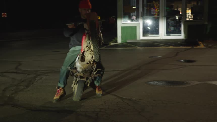 A man wearing a black shirt red cap and green pants is seen from behind doing a wheelie on a red and white dirt bike in a dark paved parking lot outside a brightly lit building at night
