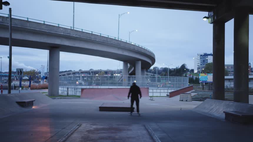 A young male adult skateboarding at an outdoor ramp in a city at dusk