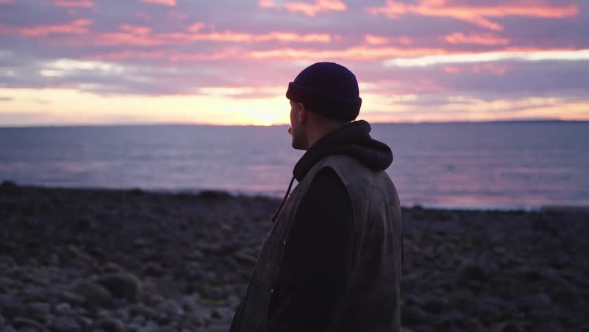 The video shows a mediumcloseup of a man standing alone on a rocky beach at sunset he is wearing a dark jacket and beanie with his back slightly turned to the camera and the sky behind him displaying soft pink and orange hues