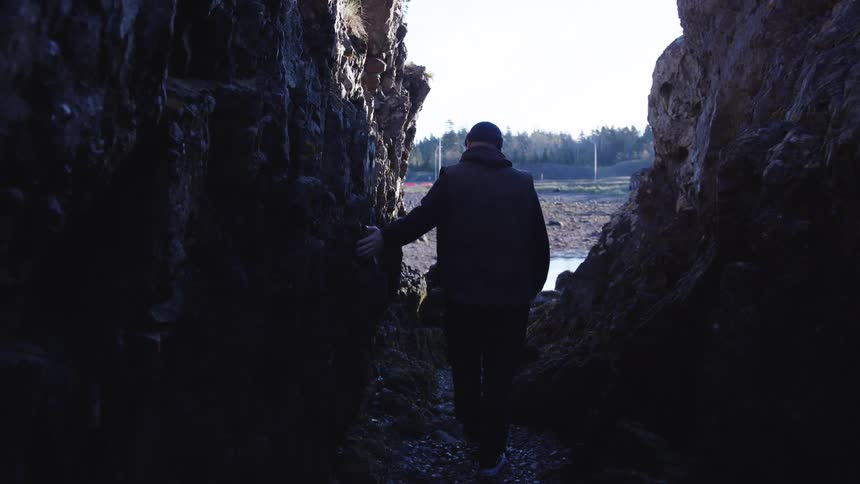 The video shows a persons silhouette emerging from the dark entrance of a rock formation or narrow cave the bright light outside reveals a coastal landscape with exposed shore possibly at low tide a body of water and a tree line in the dista