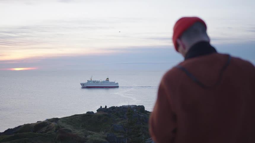 Person stands on a cliff overlooking the ocean as a ferry sails at sunset