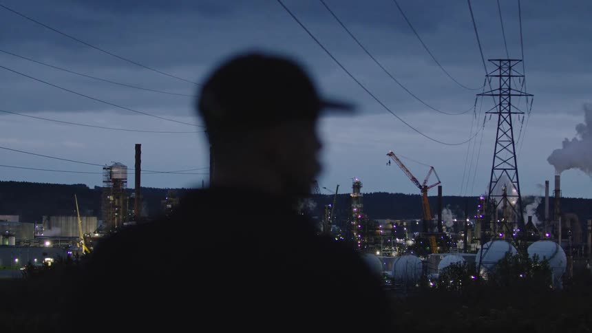 The video shows an outoffocus silhouette of a person wearing a baseball cap against a background of an oil refinery or industrial complex at dusk the brightly lit factory features tall chimneys storage tanks and a large power pylon with