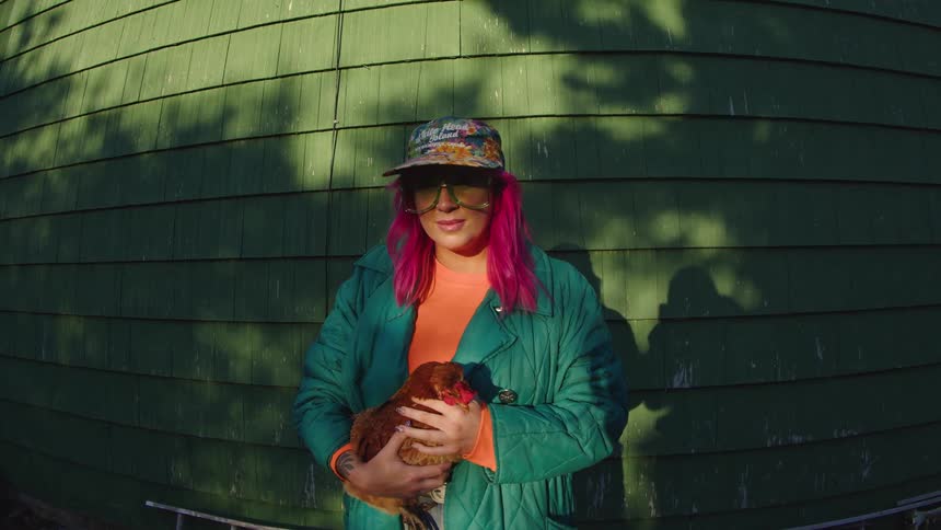 Person with pink hair holds a chicken while smiling under sunlight