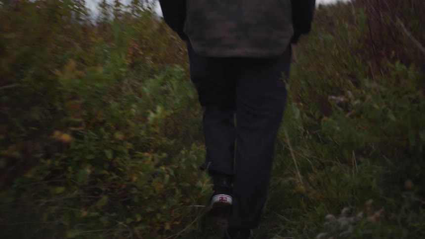 A view from behind shows a person wearing dark pants and black and white shoes walking along a narrow overgrown grass path in a field