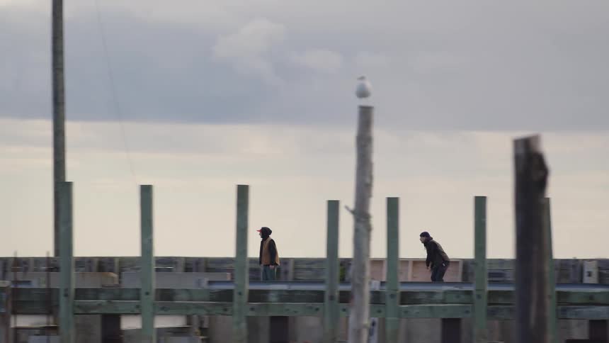 Two young boys skateboard across a bridge on a cloudy day both wearing jackets as they glide through the cool air