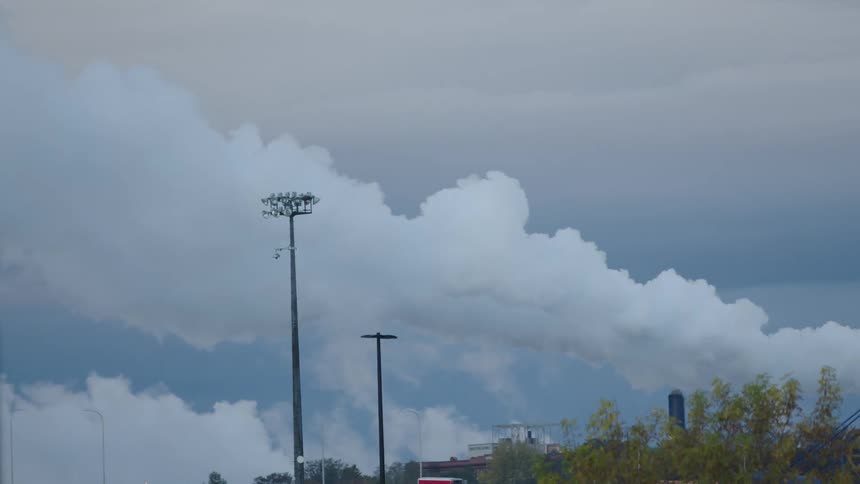 A large white cloud of smoke billows from a short stack and mixes with the low sky clouds positioned behind a tall metal light structure and a row of trees in an industrial area