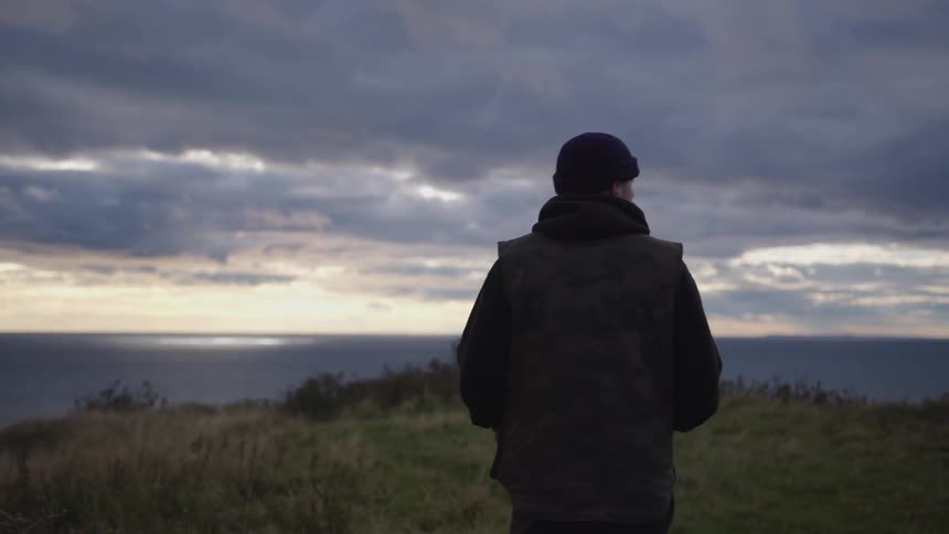 The video shows a man from behind wearing a dark jacket and a beanie standing on a grassy hill overlooking a body of water he begins to slowly walk away from the camera toward the edge of the slope gazing out at the cloudy sky and ocean ho