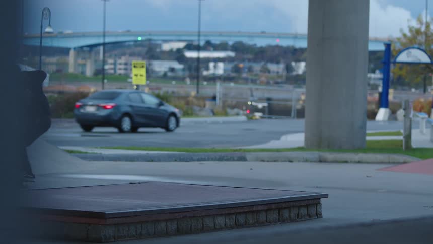 A person in dark clothing skates across a concrete skatepark performs a jumping trick over a low rectangular platform lands and continues skating toward a moving car in the background with a large bridge structure at distance