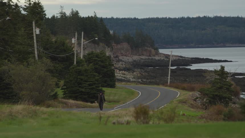 The video is a wide shot of a man walking along a winding paved road that runs parallel to a rocky shoreline and a bay the scene is backed by a densely forested hillside under an overcast sky suggesting a remote coastal location