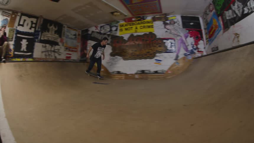 The video is a lowangle closeup shot of a person riding a skateboard up the side of a ramp inside an indoor skate parkthe walls of the park are covered in stickers and graffiti giving the space a raw urban feel