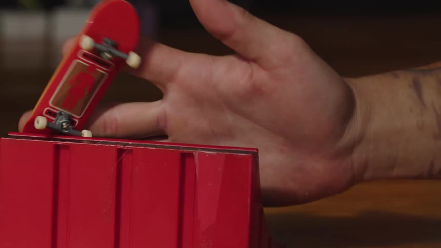 A persons hand performs a trick with a red fingerboard skateboard on a red mini ramp