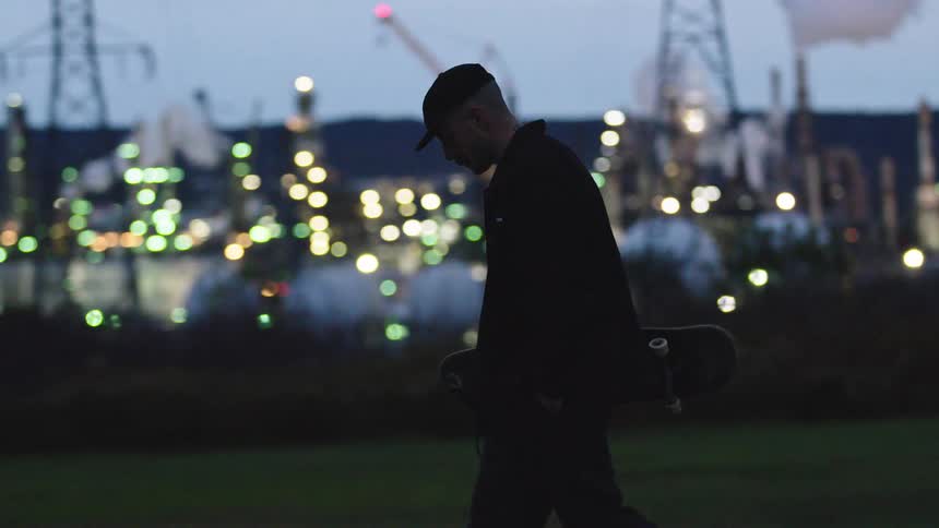 Man holds a skateboard and watches an illuminated industrial area at dusk