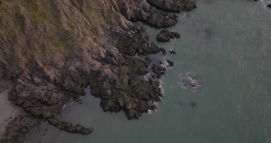 A highangle camera captures dark rough rocky cliffs with patches of vegetation on one side meeting a body of dark green water where white splashes occur