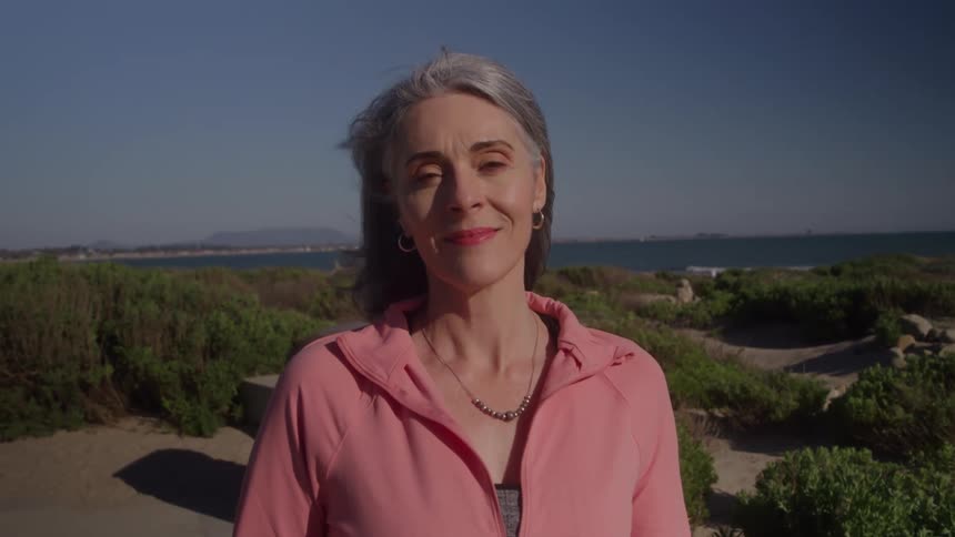 A middleaged woman with gray hair stands outdoors on a sunny day wearing a pink jacket and a necklace surrounded by grasses