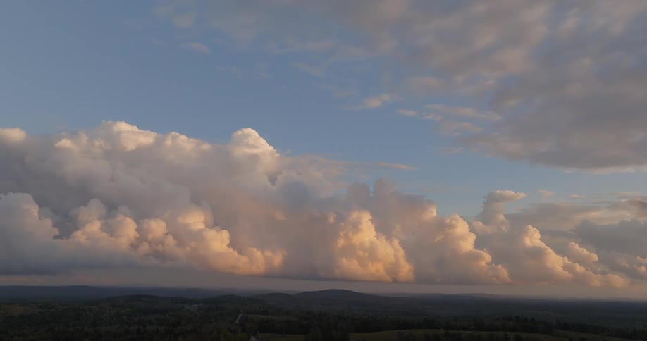 A wide elevated view captures massive cumulus clouds illuminated by warm sunset light from below towering over a dark rolling landscape of forests and hills beneath a light blue and cloudy sky