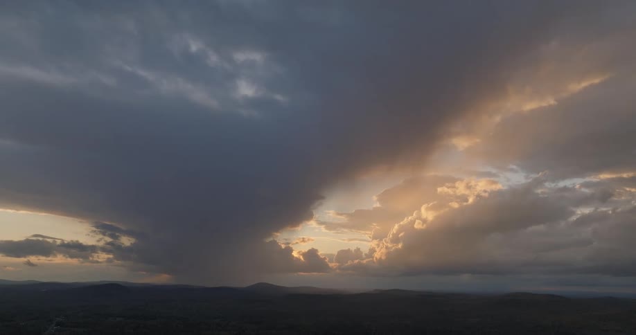Dense clouds cover part of the sky as golden light radiates in the background over forested mountain range