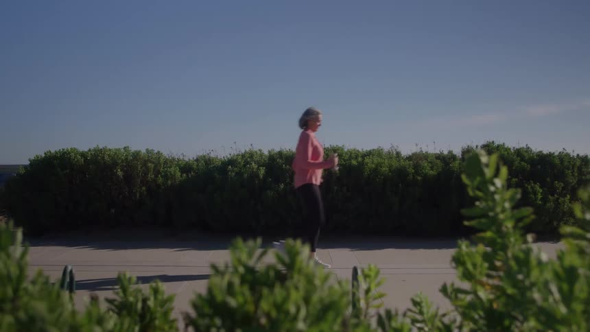 The video is a sideview shot of a woman with blonde hair walking briskly or jogging from left to right on a sidewalk she is wearing a pink top and dark leggings and passes behind a low wellmanicured hedge under a clear blue sky