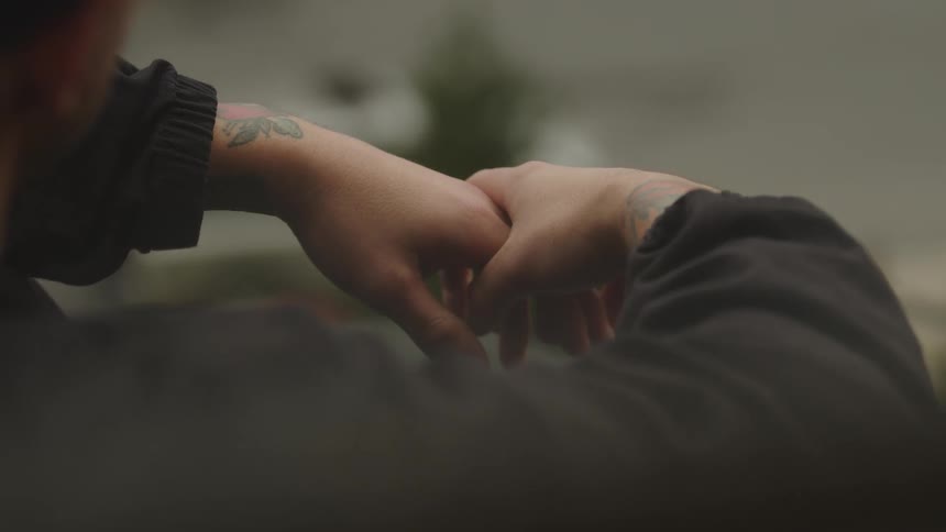 Two tattooed forearms and hands emerging from dark sleeves gently reach out and grasp each other in the foreground with a blurred background