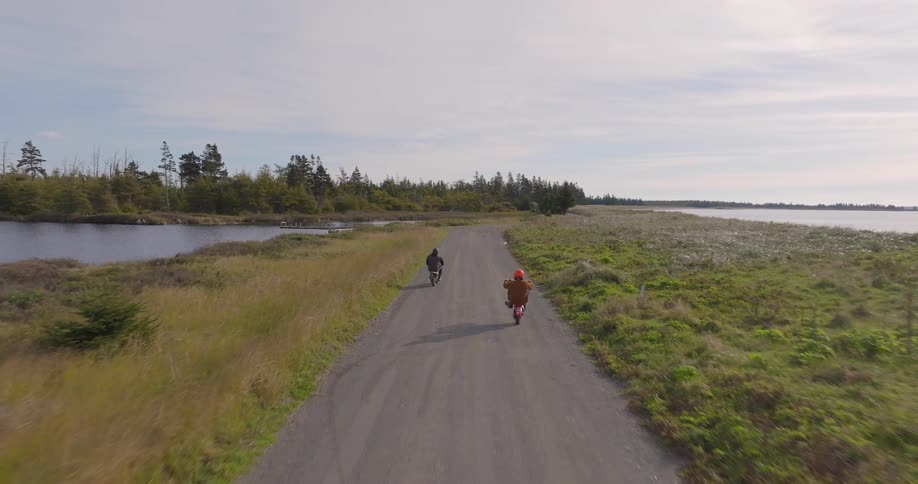 Two motorcyclists riding along a narrow road surrounded by vegetation and water
