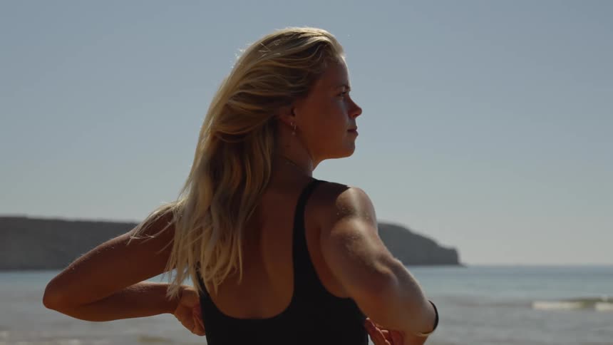 Close up portrait of a young blonde woman adjusting her swimwear while standing outdoors at the beach during daytime