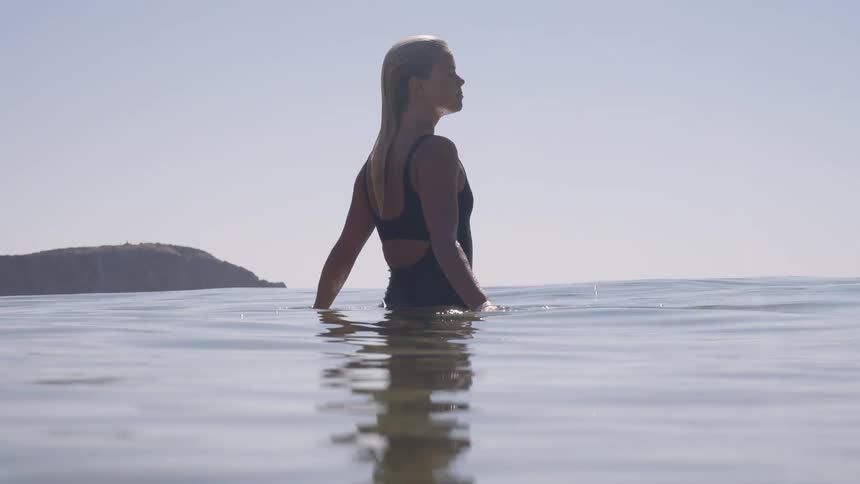 A person stands chest deep in calm ocean water near the shore gazing out toward a rocky headland under a clear sky