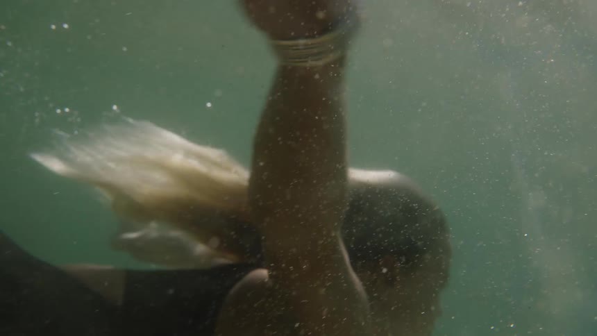 Lighthaired woman swims underwater in aquatic environment lit with soft shadows and fluid motion