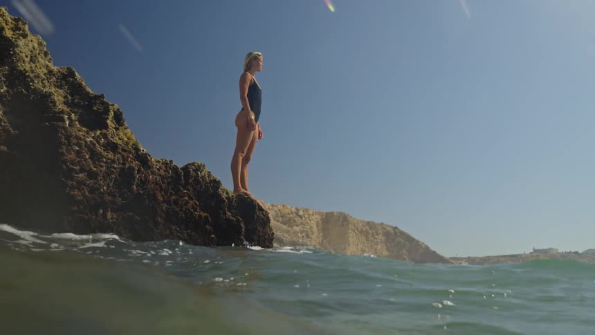 A person stands on a rocky outcrop near the ocean the person wears a dark swimsuit and looks out waves crash below the rock water splashes upward and tall cliffs line the background under a blue sky