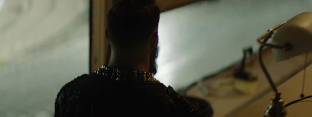A person with a beard and dark clothing featuring a studded collar walks past an opening moves into an indoor area turns their back to the camera and walks toward a table with a television monitor displaying static