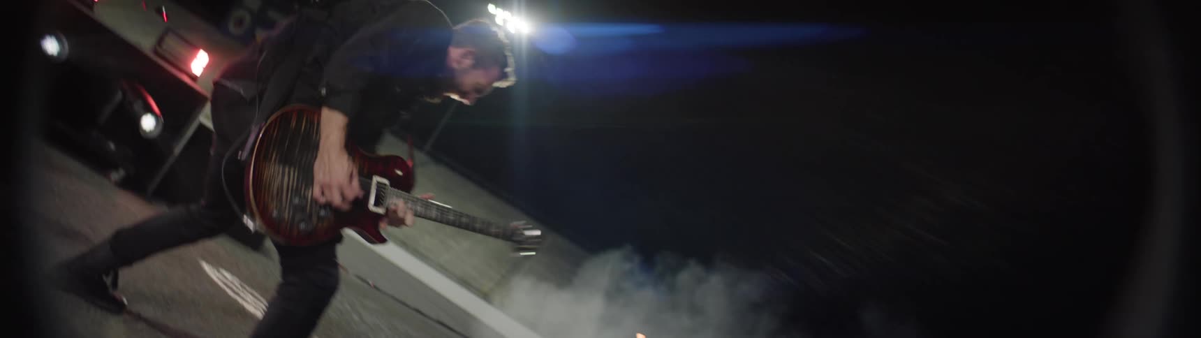 The video features an extreme lowangle dynamic shot of a musician vigorously playing an electric guitar on a stage during a night performance the scene is dark dominated by bright stage lights and a cloud of fog or smoke swirling near the