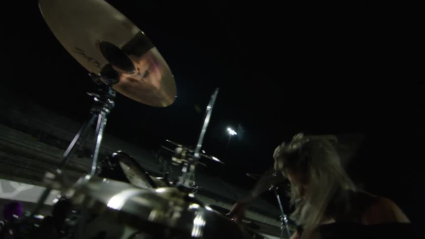 A blonde woman wearing a dark off the shoulder top energetically plays a drum set with visible cymbals on an outdoor stage at night with bright stadium lights shining onto the dark field