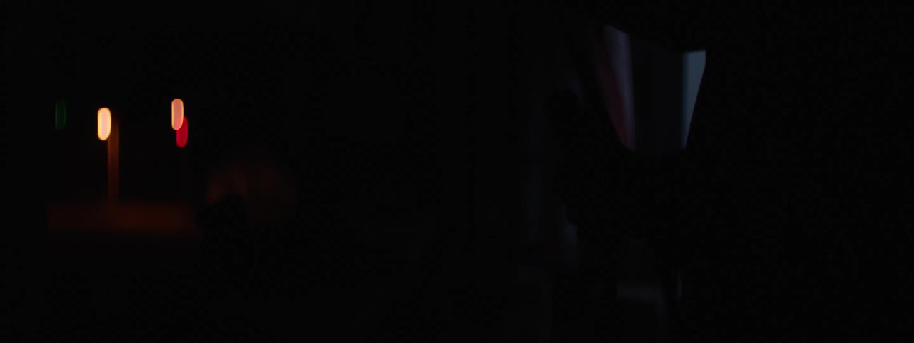 A persons hands grip a microphone stand in a dark room with a small glowing white screen from a television monitor in the background