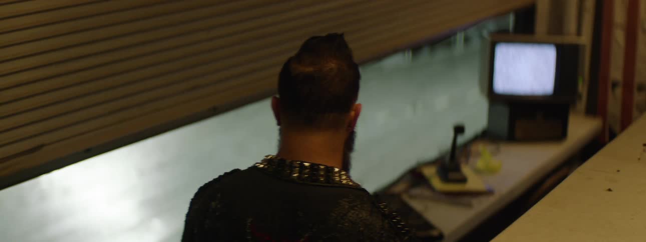 A high angle medium view from behind shows a man with a beard and hair pulled back wearing a black shirt with gold pyramid studs on the collar looking toward a small static filled television monitor in a room