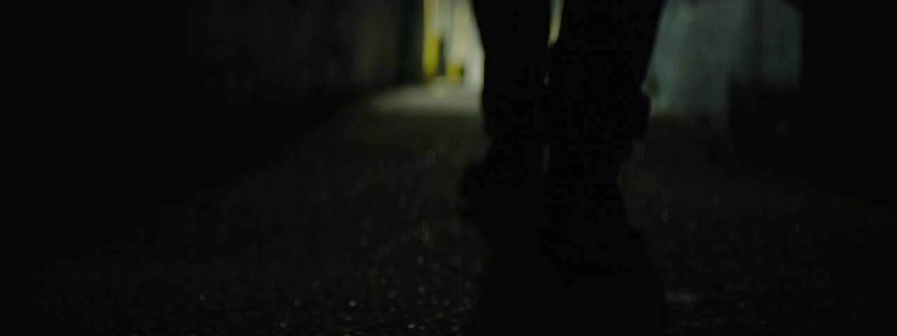 A young man walking along a dark narrow corridor as he moves toward the distant light the camera follows and reveals a black jacket and the corridor opens up to an outdoor area with bright lights and a fence