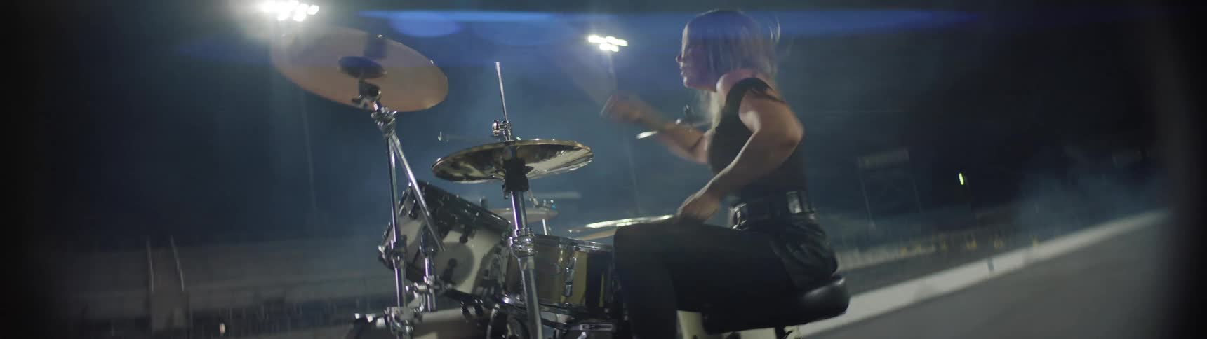 A dynamic low angle shot captures a blonde woman in a dark outfit aggressively playing a drum set and cymbals on a dark outdoor stage intensely backlit by bright stadium lights with visible lens flare