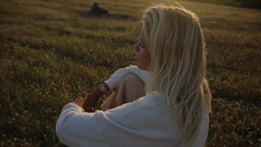 A young woman with blonde hair sits on a grassland wearing a sweatshirt looking around as the wind lifts a bit of her hair