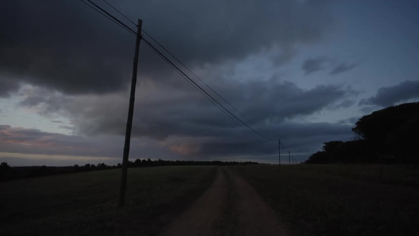A rural dirt road extends into the distance between dark fields flanked by utility poles under a heavy twilight sky filled with dark clouds