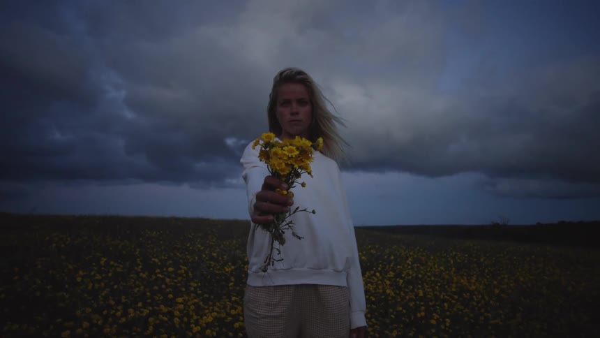 A person stands in a field of yellow flowers beneath a cloudy sky holding a small bouquet