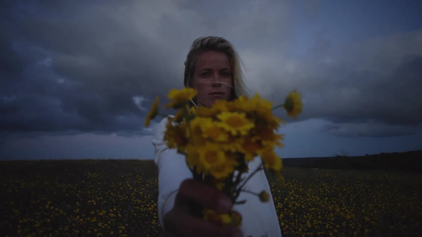 A young woman stands in a field of yellow flowers and holds a bouquet of the same flowers directly toward the camera and the background shows a cloudy dark sky over a flat landscape