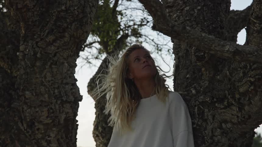 A close low angle shot shows a blonde woman wearing a white sweatshirt standing between two large dark tree trunks looking up at the sky as the wind gently blows her hair
