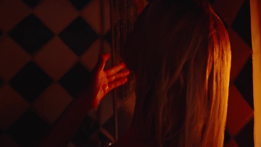 A woman with wet blonde hair stands under a shower lifting a hand to the stream in warm orange light against black and white tiles