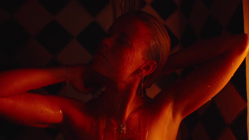 A person stands under a stream of water in a dimly lit enclosure runs hands through hair and over the head and water flows down the front of the body against a background of patterned tiles