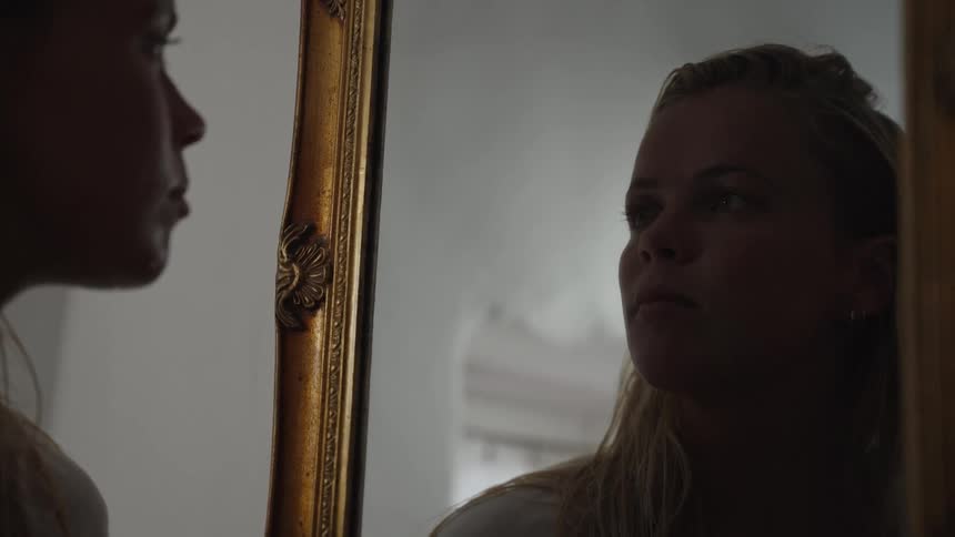 A woman looks into a tall gold framed mirror in dim light