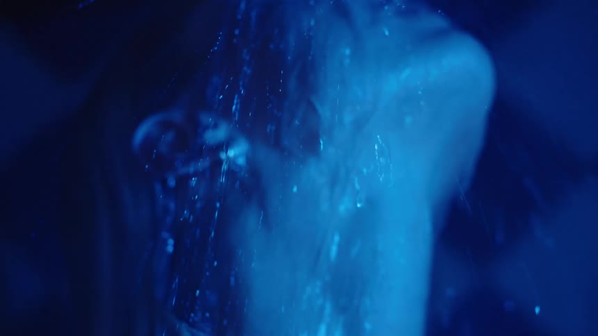 A close view shows a woman with blonde hair wearing a silver necklace in a dark shower tilting her face upward as streams of water run down her body under bright blue lighting