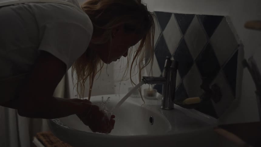 A blonde woman wearing a white shirt turns on a faucet and splashes water on her face in a dimly lit bathroom with blue and white diamond patterned tiles then looks at her reflection in a gold framed mirror