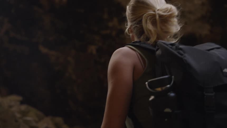 A young woman with blonde hair hikes across rocky terrain at midday carrying a backpack