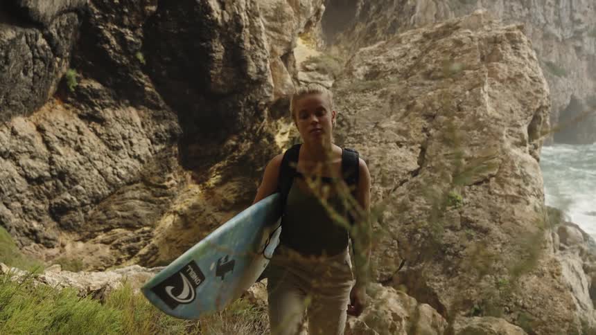 Woman crosses rocky terrain carrying a surfboard and backpack near a coastal area