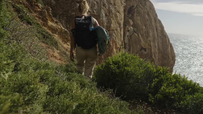 A person walks down a dirt path carrying a surfboard and a backpack moving past large rock formations dense green bushes line the trail and the ocean is visible in the background under the bright sky