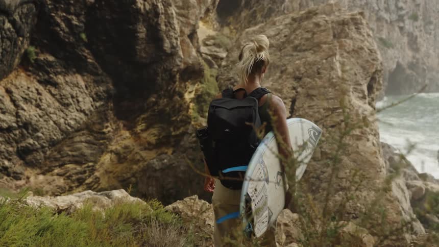 Woman with a surfboard and backpack climbing among rocks toward the sea